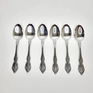 May include: Six silver spoons arranged in a row. Each spoon has a rounded bowl and a decorative handle with an ornate floral design. The spoons are made of a shiny, reflective metal, likely stainless steel.