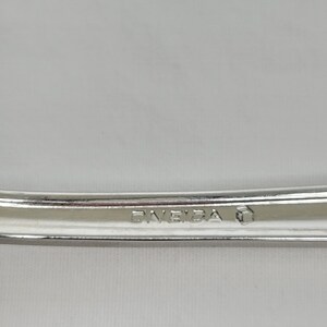 May include: Close-up of a silver utensil with the word "ONEIDA" stamped on it. The utensil has a curved design and a shiny finish. The background is a solid, light colour.