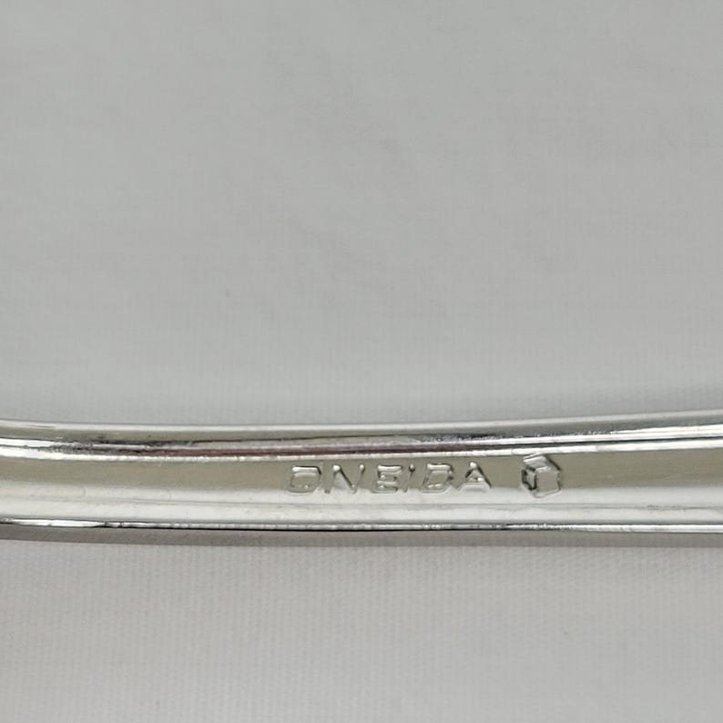 May include: Close-up of a silver utensil with the word "ONEIDA" stamped on it. The utensil has a curved design and a shiny finish. The background is a solid, light colour.