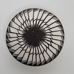 May include: A black and white pottery plate with a geometric design. The plate has a central hole and a swirling pattern of white lines on a black background.