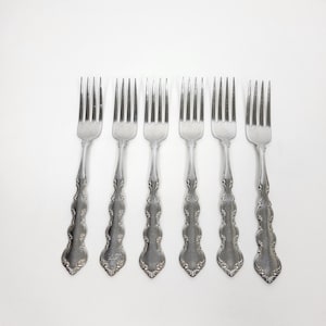 May include: A set of six silver-toned metal forks arranged in a row. Each fork features four tines and a handle with a decorative embossed scroll pattern along the edges, tapering to a rounded base. The finish is smooth and reflective.
