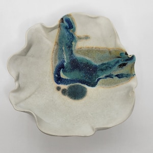 May include: A ceramic plate with a ruffled edge and a white base. The plate features a blue and teal abstract design in the center. The design has a flowing, organic shape with darker blue accents.