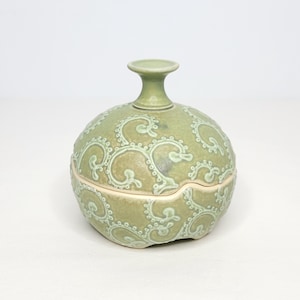 May include: A light green ceramic jar with a lid, featuring a decorative swirl pattern. The jar has a rounded body and a small, cylindrical handle on top. The lid fits snugly, and the jar is likely used for storage.