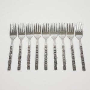 May include: Nine stainless steel forks with a decorative floral pattern on the handles. The forks are arranged in a row against a white background, showcasing their tines and detailed design.