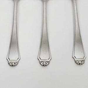 May include: Close-up of three silver spoons. Each spoon has a long, slender handle that widens towards the top. The spoon bowls have a decorative, scalloped edge with a floral design. The spoons are arranged vertically.