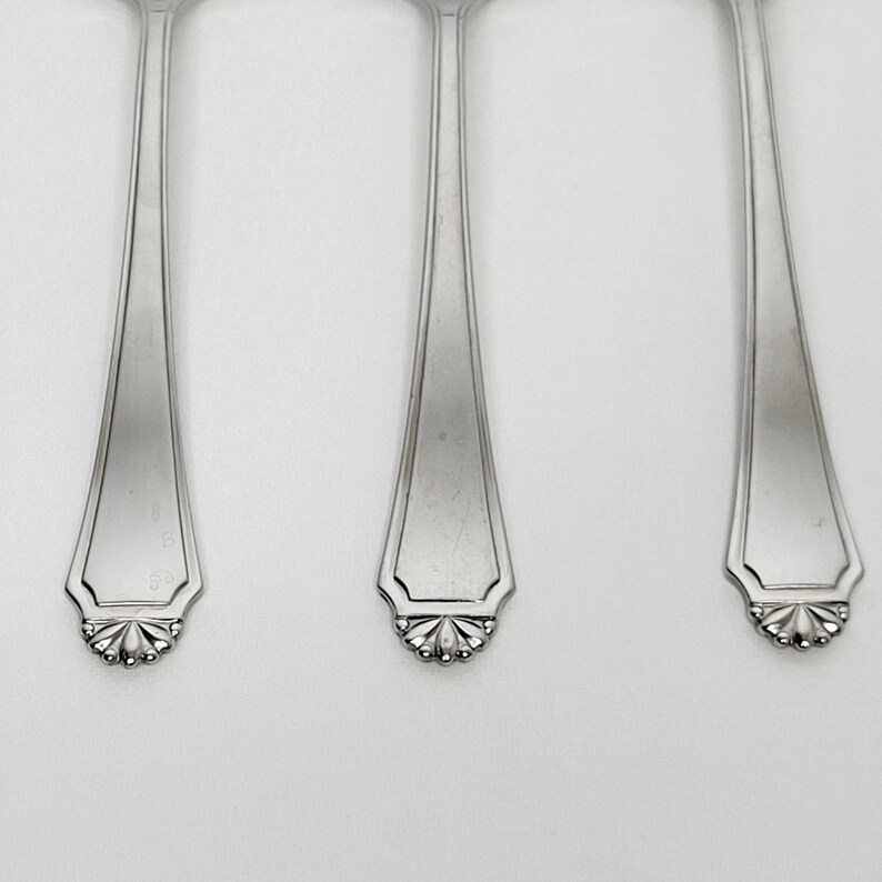 May include: Close-up of three silver spoons. Each spoon has a long, slender handle that widens towards the top. The spoon bowls have a decorative, scalloped edge with a floral design. The spoons are arranged vertically.