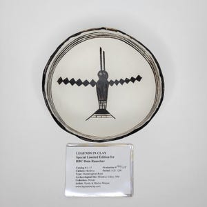 May include: A white ceramic bowl with a black hummingbird design in the center. The bowl has a black rim with two concentric black lines. The text "LEGENDS IN CLAY" is visible on a label below the bowl.
