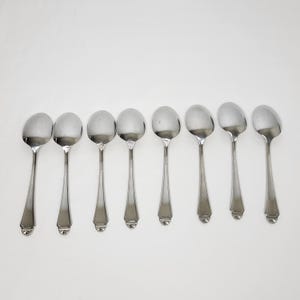 May include: Eight silver soup spoons on a white backdrop. Each spoon features a rounded bowl and a long, slender handle with a decorative base. The spoons are evenly spaced, reflecting light. Ideal for serving soup or other dishes.