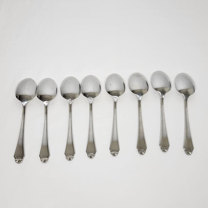 May include: Eight silver soup spoons on a white backdrop. Each spoon features a rounded bowl and a long, slender handle with a decorative base. The spoons are evenly spaced, reflecting light. Ideal for serving soup or other dishes.