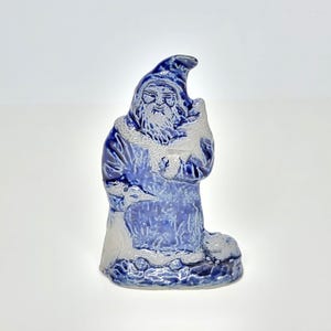 May include: A ceramic figurine of a bearded figure wearing a hooded robe, finished in a glossy blue glaze with unglazed white textured accents on the hood, beard, cuffs, and base. The figure holds a small bird and stands on a rocky base.