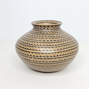 May include: A round, beige and black ceramic vase with a wide body and a narrow neck. The vase is decorated with repeating geometric patterns in black against a beige background. The patterns create horizontal bands around the vase.