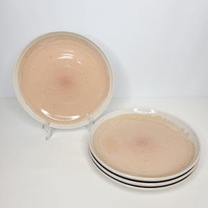 May include: A set of four round ceramic plates featuring a soft peach-colored center with concentric circular ridges and a speckled finish. The plates have a wide, smooth, off-white rim and a glossy glaze, presented as one upright plate and a stack of three.