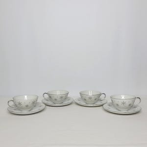 May include: Four white teacups and saucers with a silver snowflake design. Each cup has a silver rim and handle, and the saucers have a matching snowflake pattern. The set is arranged on a white surface.