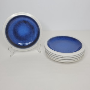 May include: A set of six ceramic plates. Each plate has a white rim and a blue center with a speckled design. One plate is displayed on a clear plastic stand. The plates are round and appear to be new.