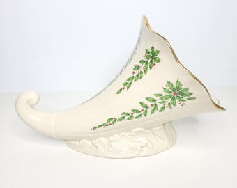 May include: A cream-coloured ceramic cornucopia vase featuring a painted green holly leaf and red berry pattern along the side. The horn-shaped vessel has a flared, wavy rim accented with a thin gold trim and rests on a sculpted base.
