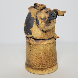 May include: A ceramic sculpture of a pig with black and brown markings. The pig is sitting on a round, brown ceramic base.