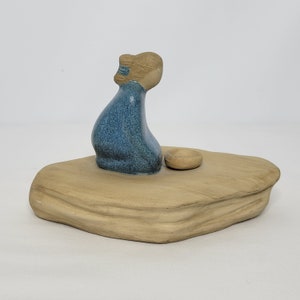 May include: A ceramic figurine of a person sitting on a beige rock with a small bowl in front of them. The person is wearing a blue robe and has brown hair.