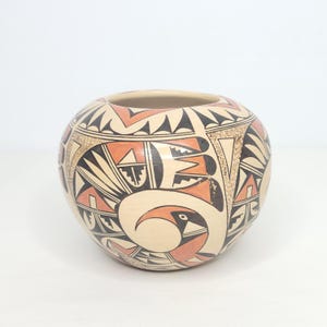 May include: A round, cream-colored ceramic pot with intricate geometric and bird designs in black, orange, and tan. The pot features a wide opening and a rounded body, showcasing traditional Native American pottery.