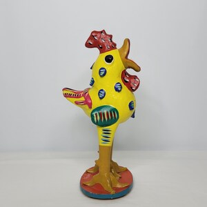 May include: A yellow ceramic rooster figurine with red, blue, and green painted details. The rooster has a yellow body with blue polka dots, a red comb, and a red wattle. The rooster's legs are painted yellow and brown, and the base is painted red, blue, and yellow.