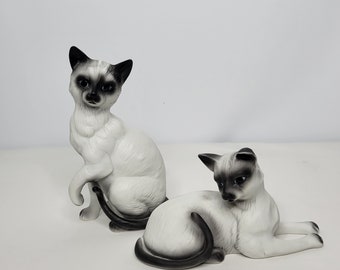 May include: Two white and black Siamese cat figurines. The cats are in a sitting position with their tails curled around their bodies. The figurines are made of porcelain and have a glossy finish.