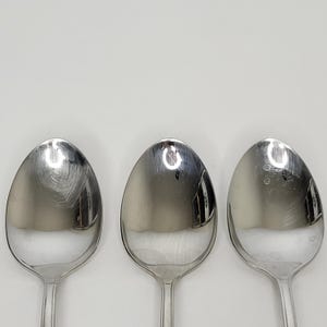 May include: Three stainless steel spoons with oval bowls and long, slender handles. The spoons are arranged vertically against a plain white background. The spoons reflect the light, creating bright highlights on their polished surfaces.