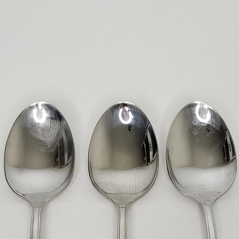 May include: Three stainless steel spoons with oval bowls and long, slender handles. The spoons are arranged vertically against a plain white background. The spoons reflect the light, creating bright highlights on their polished surfaces.