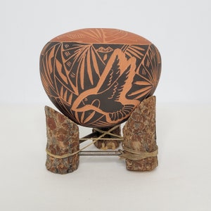May include: A hand-painted terracotta pottery bowl with a black and brown design of a bird in flight. The bowl is on a wooden stand with a metal wire frame.
