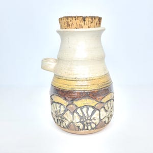 May include: A handcrafted ceramic bottle with a cork stopper. The bottle features a cream-colored top, a yellow and brown band, and a floral pattern at the base. It has a small handle on the side. The bottle is approximately 7 inches tall.