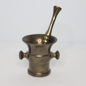 May include: A brass mortar and pestle set. The mortar is a small, round bowl with two handles. The pestle is a long, cylindrical rod with a rounded end.