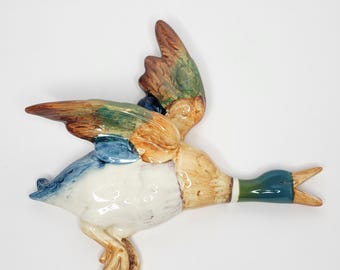 May include: A ceramic duck figurine in flight, featuring a detailed design with brown, green, blue, and white colors. The duck has outstretched wings and an open beak, showcasing its realistic features. This decorative piece is ideal for home decor.