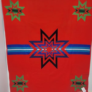 May include: Red wool blanket with a geometric pattern of green, blue, and black stars. The blanket has a white label with black text.