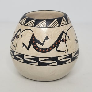 May include: A small, white ceramic pot with black and orange painted designs of lizards. The pot has a rounded shape and a wide opening.