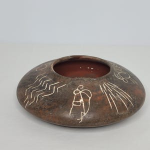 May include: A brown ceramic bowl with white painted designs. The bowl has a wide, flat base and a rounded rim. The designs include geometric patterns and a stylized human figure.