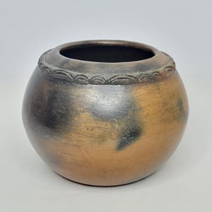 May include: A handcrafted, round ceramic pot with a wide opening. The pot features a dark brown interior and a textured rim with a repeating pattern. The exterior has a gradient of colors, from light brown to dark brown and black.