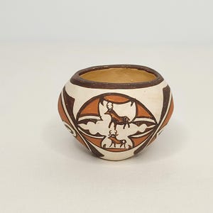May include: A small, round, hand-painted ceramic pot with a cream-colored base and brown and orange geometric designs. The pot features two deer figures. The rim is brown, and the interior is a light tan color.