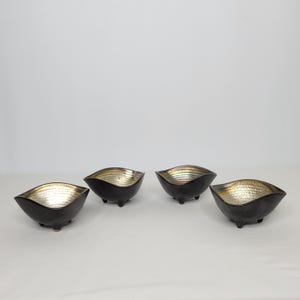 May include: Four black ceramic bowls with a textured, iridescent gold interior. The bowls have a unique, wavy shape and sit on three small feet. The bowls are arranged on a white surface.