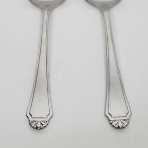 May include: Two silver spoons with a decorative floral design at the base of the handle. The spoons have a classic, elegant shape and are set against a plain white background. The spoons are made of a shiny, reflective metal.