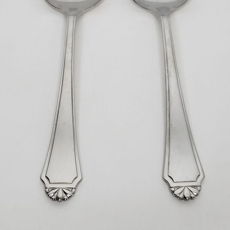 May include: Two silver spoons with a decorative floral design at the base of the handle. The spoons have a classic, elegant shape and are set against a plain white background. The spoons are made of a shiny, reflective metal.