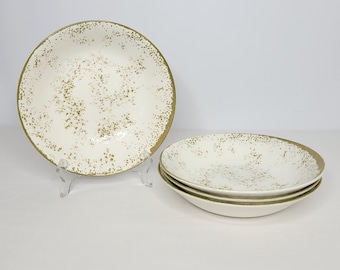 May include: A set of four off-white ceramic bowls with a speckled gold pattern. The bowls have a gold rim and are displayed on a white surface. One bowl is on a clear stand, and three are stacked.