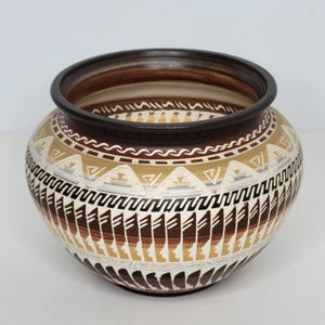 May include: A round, handcrafted ceramic pot with a dark brown rim and a detailed geometric pattern. The design incorporates shades of brown, beige, and white, creating a visually appealing, earthy aesthetic. The pot's shape is wide and rounded, suitable for decorative use.