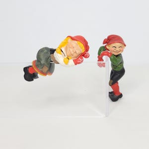 May include: Two whimsical gnome figurines. One is lying down, wearing a red hat, yellow hair, and green pants. The other gnome is standing, wearing a red hat, green shirt, and black pants. Both have rosy cheeks and are smiling.