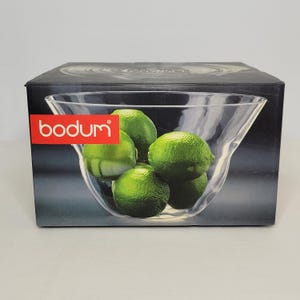 May include: A clear glass bowl filled with green limes, presented in a black box with the brand name "bodum" in white letters on a red rectangle. The bowl is displayed on the box, showcasing the product.