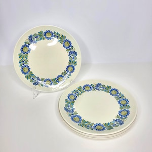 May include: A set of three round ceramic plates featuring a cream-colored base and a decorative border pattern of blue and green stylized flowers and leaves arranged in a circular wreath design around the center of each plate.