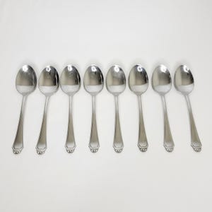 May include: Seven stainless steel spoons on a white background. Each spoon has a rounded bowl and a long, slender handle. The handles feature a decorative design at the base. The spoons are evenly spaced and reflect the light.