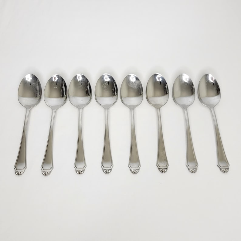 May include: Seven stainless steel spoons on a white background. Each spoon has a rounded bowl and a long, slender handle. The handles feature a decorative design at the base. The spoons are evenly spaced and reflect the light.