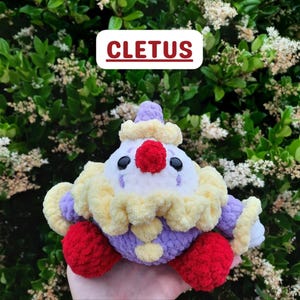 以下が含まれることがあります： CLETUSという名前の、かぎ針編みのピエロのぬいぐるみ。白い顔、鮮やかな赤い鼻、黒い目、黄色いフリルの襟が特徴です。紫色の糸で作られた体に、赤い足と紫色の袖が付いており、全体的に柔らかく質感のある毛糸素材で仕上げられています。
