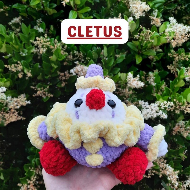 以下が含まれることがあります： CLETUSという名前の、かぎ針編みのピエロのぬいぐるみ。白い顔、鮮やかな赤い鼻、黒い目、黄色いフリルの襟が特徴です。紫色の糸で作られた体に、赤い足と紫色の袖が付いており、全体的に柔らかく質感のある毛糸素材で仕上げられています。