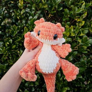 Peut inclure: Une peluche en crochet faite à la main, représentant un lézard orange et blanc, avec des yeux noirs. La peluche a un ventre blanc et un aspect texturé et flou. Le lézard est tenu devant un fond de feuillage vert.