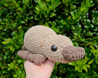 Puede incluir: Un juguete de ornitorrinco tejido a ganchillo con hilo beige suave y texturizado. La criatura presenta un pico marrón oscuro, pequeños ojos de seguridad de plástico negro y cuatro patas palmeadas. El peluche tiene un cuerpo redondeado y una cola plana, elaborado con un patrón de puntos denso y consistente en todo el diseño.