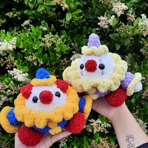 K&ouml;nnte beinhalten: Zwei geh&auml;kelte Stofftiere in Form von Clowns. Der eine ist gelb, wei&szlig; und lila mit einer roten Nase. Der andere ist rot, gelb und blau mit einer roten Nase.